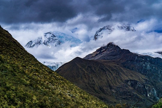 Snow-capped peaks behind a green slope under dark clouds