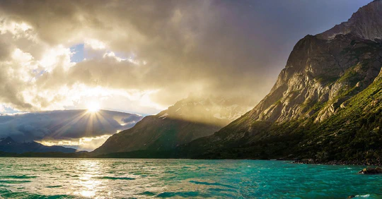 The sun breaking through clouds over a turquoise lake and mountains