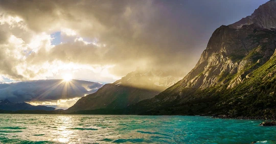 The sun breaking through clouds over a turquoise lake and mountains