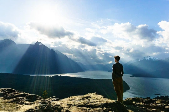 A person on a rocky ledge watching sunbeams fall on mountains and a lake