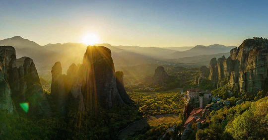 Sunset over tall rock pillars and a valley, with red-roofed buildings on a rock